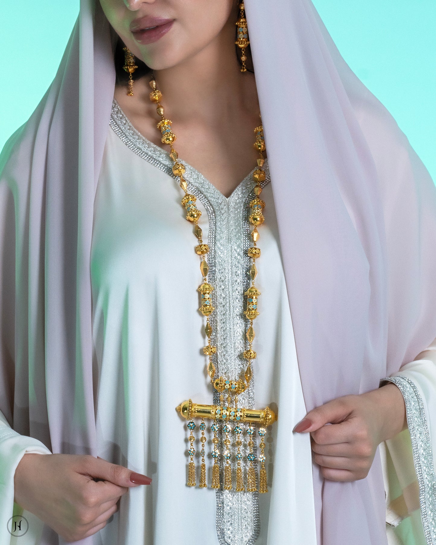 This 21K yellow gold traditional Bahraini long necklace set features a mezuzah-inspired design in mina work, adorned with bead and small flower hangings. The rope-length necklace fastens with a fish-eye clasp and is accompanied by matching drop earrings and a ring.
A traditional Khaleeji jewelry piece, perfect for bridal, wedding, or special occasions. This elegant Arabic design pairs beautifully with abayas.