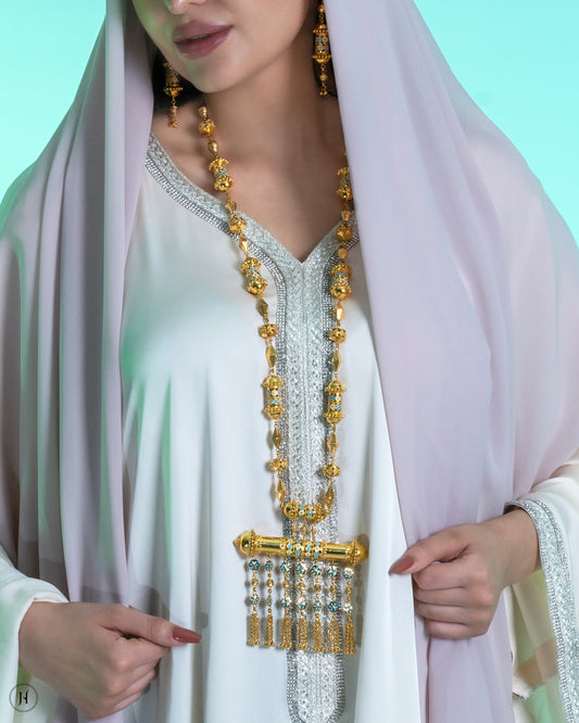 This 21K yellow gold traditional Bahraini long necklace set features a mezuzah-inspired design in mina work, adorned with bead and small flower hangings. The rope-length necklace fastens with a fish-eye clasp and is accompanied by matching drop earrings and a ring.
A traditional Khaleeji jewelry piece, perfect for bridal, wedding, or special occasions. This elegant Arabic design pairs beautifully with abayas.